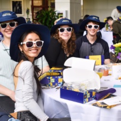 A group of smiling young adults sits at a table, each wearing white sunglasses and matching blue hats. 的y are surrounded by gift boxes, pamphlets, and floral arrangements. 的 setting appears to be a casual event or gathering.