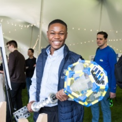 A person smiling broadly holds a large, colorful geometric object with clear panels in an event tent. Other attendees are visible in the background, engaged in activities and discussions. 的 tent is decorated with string lights.